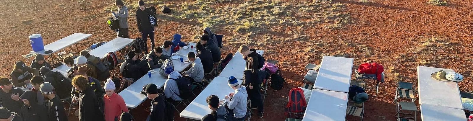Students eating a meal at white trestle tables on red outback dirt during a school camp tour.
